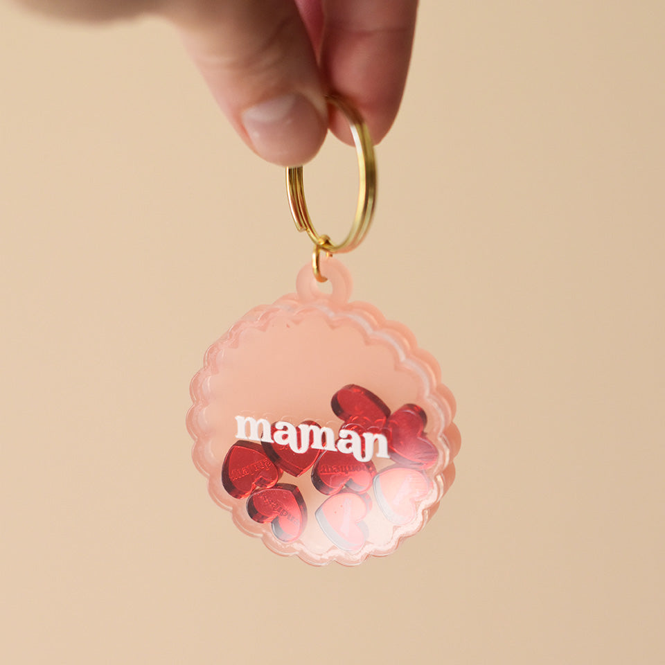 Porte-clés en plexiglass transparent et coloré rose sur anneaux dorés. Il a une forme de cercle festonné et contient 8 mini découpes en forme de cœur rouge qui bougent librement à l’intérieur. Ces découpes sont gravées de mots associés à la maternités « tendresse, amour, bisous.. »